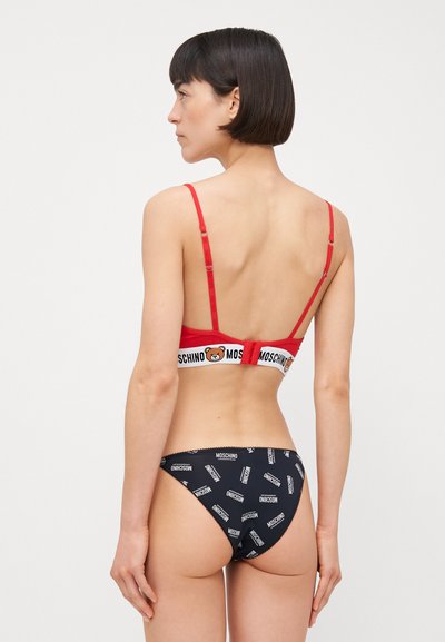 Sarkana bralette ar logo jostas un regulējamiem siksniņām, savienota ar melnajiem bikini apakšbiksēm ar atkārtotu baltu zīmola tekstu un zemu vidukli.