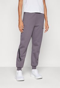 Grå joggebukser laget av mykt stoff, med sidelommer med glidelås og svarte detaljer. Tapered fit med ribbestrikkede mansjetter ved ankelen.