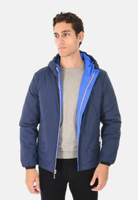 Giacca imbottita da uomo con esterno blu navy, fodera interna blu a contrasto e chiusura con zip. Include un cappuccio e polsini a coste.