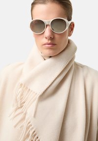 Bílý šátek s třásněmi přehoděný přes krk, doplněný kulatými světle zbarvenými slunečními brýlemi a hladkým, elegantním pozadím.