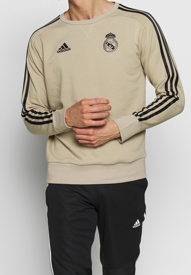 Felpa beige Adidas con strisce nere e logo del Real Madrid, abbinata a pantaloni da jogging neri Adidas con strisce bianche.