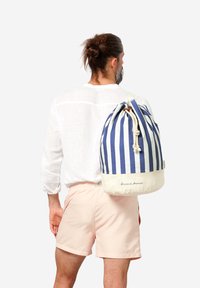 Mochila de cuerdas a rayas en lona azul y blanca, con una base crema contrastante y un cierre decorativo de cuerda.