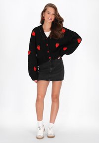 Kvinna i svart kofta med röda jordgubbsmönster, svart denimminikjol, vita sneakers och röda körsbärsörhängen, stående och leende.