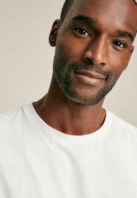 Valge puuvillane T-särk ümmarguse kaelusega, siledate tekstuuriga, esitatud mehemudeli peal, rõhutades näojooni ja nahatooni.