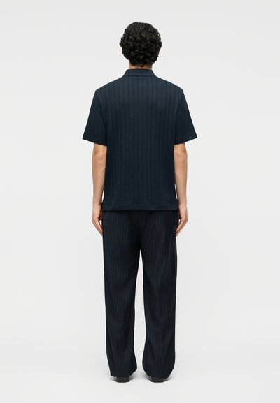 Άνδρας με σγουρά μαλλιά, στεκόμενος με την πλάτη προς τα εμπρός, φορώντας ένα σκουρόχρωμο μπλε πουκάμισο με λεπτές ρίγες και κοντά μανίκια, καθώς και ένα ζευγάρι χαλαρά παντελόνια που ταιριάζουν.