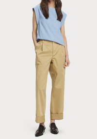 Lyseblå ærmeløs top, khaki plisserede bukser med ombrettede manchetter og sorte slip-on sko med præget design. Glatte, afslappede stofteksturer.