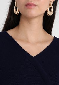 Top a scollo a V navy in tessuto maglia testurizzato, abbinato a grandi orecchini a cerchio d'oro. I dettagli si concentrano sul scollo e sul design degli orecchini.