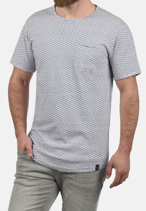 Uomo che indossa una t-shirt a maniche corte bianca e nera a righe con taschino sul petto e pantaloni grigio chiaro, mano in tasca.