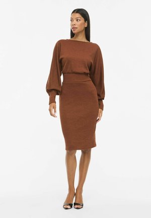 Modèle portant une tenue brun rouille composée d’un haut ample à manches longues et d’une jupe ajustée jusqu’aux genoux, assortie à des escarpins noirs à bout ouvert.