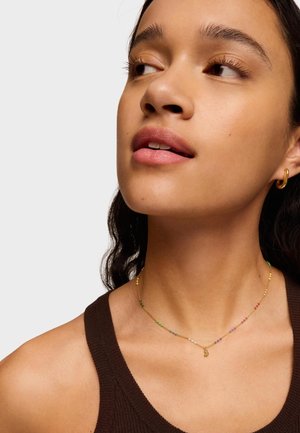 Collar de cadena de oro con cuentas multicolores, que presenta un pequeño charm. El modelo lleva una blusa sin mangas de color marrón, mostrando claramente la joyería.