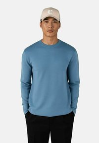 Blå genser med rund hals i en glatt strikket tekstur, kombinert med en beige caps med logo foran. Modellen har på seg svarte bukser.