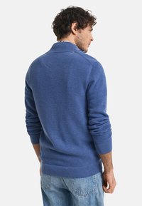 Mann, der seitlich steht, trägt einen strukturierten blauen Pullover mit gerippten Bündchen und Kragen, kombiniert mit hellblauen Jeans.