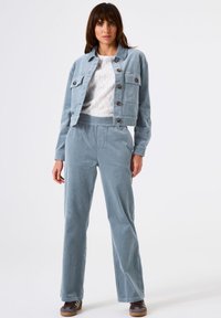 Frau trägt eine hellblaue Cordjacke und -hose, ein weißes Stricktop und braune Sneakers und steht vor einem schlichten weißen Hintergrund.