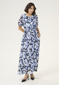 Robe maxi bleu clair avec un motif floral noir et blanc. Comprend des manches courtes bouffantes, une taille élastique et des poches latérales. Associée à des talons noirs.