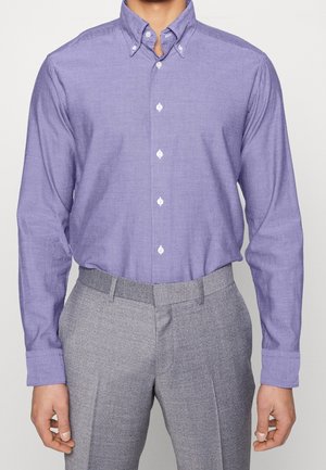 Man die een dichtgeknoopte lavendelkleurige lange mouw blouse draagt, ingestopt in een lichtgrijze broek met een hoge taille, staande tegen een effen achtergrond.