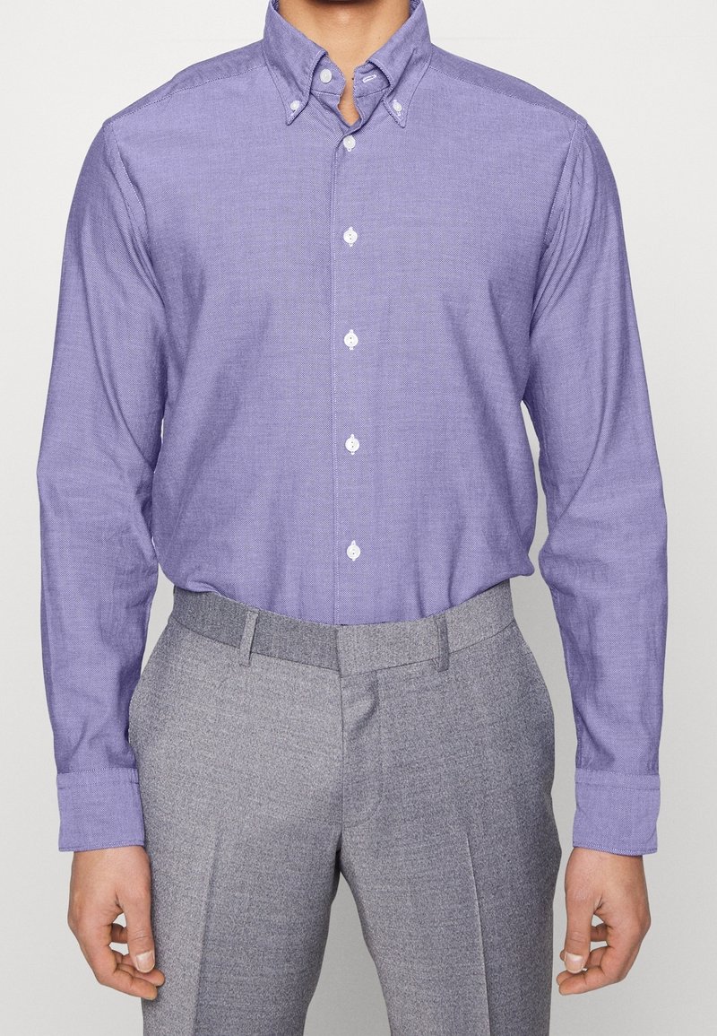 Homme portant une chemise lavande à manches longues boutonnée et rentrée dans un pantalon de costume gris clair taille haute, debout devant un fond uni.