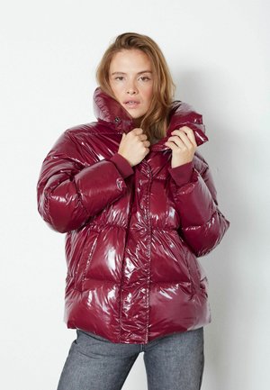 Geacă puffer burgundy cu un finisaj lucios, cu guler înalt, design quilted și manșete elastice, purtată cu pantaloni gri.