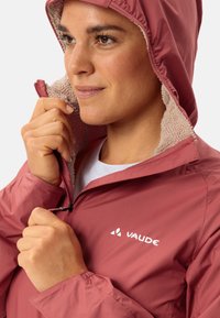 Helle rosa Kapuzenjacke mit einem weichen Fleece-Innenfutter, ausgestattet mit einem Reißverschluss und einem Logo auf der Brust. Leichter Stoff mit einer glatten Textur.