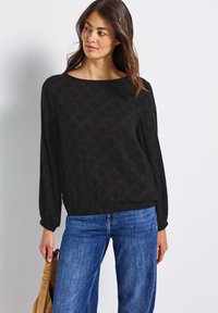 Zwarte blouse met lange mouwen en een geometrisch bruin patroon, ronde halslijn en een losse fit; gecombineerd met blauwe spijkerbroeken en een bruine tas.