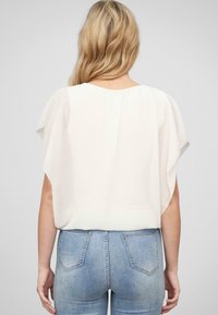 Blusa bianca trasparente con maniche corte e ampie, abbinata a jeans aderenti azzurri. Il tessuto sembra leggero e leggermente traslucido.