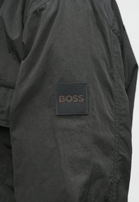 Black nylon jacket featuring a textured surface, logo patch in black and brown on the sleeve, showcasing a modern, functional design.