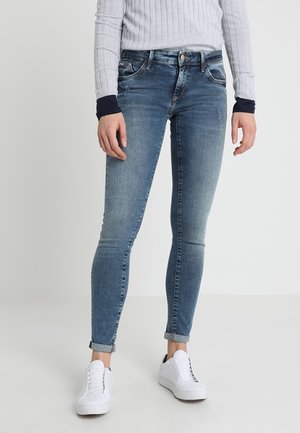 Slim fit -tyyliset siniset farkut käännettynä nilkoista, yhdistettynä valkoisiin lenkkareihin ja valkoiseen ribbineulottuun pitkähihainen toppiin, jossa on mustat kalvosimet.