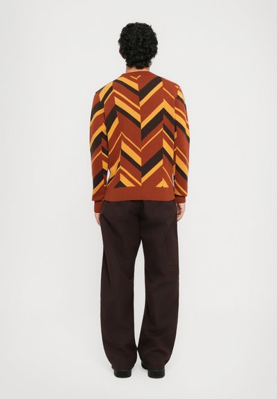Man som bär en sweater med zigzag-mönster i rost, gult och svart, samt lösa mörkbruna byxor, stående med ryggen mot kameran mot en vit bakgrund.