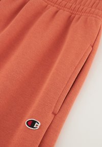Pantalons de survêtement en coton orange avec une taille élastique, des poches latérales et un petit logo brodé sur la jambe gauche. Texture douce.