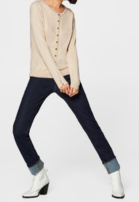 Beige long-sleeve button-up sweater, dark blue jeans with rolled cuffs, and white ankle boots with a block heel. Simple, casual outfit.