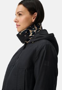 Vrouw in zwarte jack met capuchon, met een hoge kraag met luipaardprint, kijkt naar links tegen een effen lichte achtergrond.
