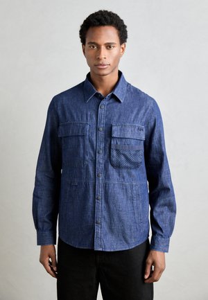 Denim blue button-up shirt with long sleeves, two chest pockets, and a mesh accent pocket. Smooth texture and curved hem.