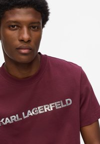 Burgundijas kokvilnas t-krekls ar sudraba metāla logotipu "KARL LAGERFELD" uz krūtīm. Apaļš kakls, īsas piedurknes, gluda tekstūra.