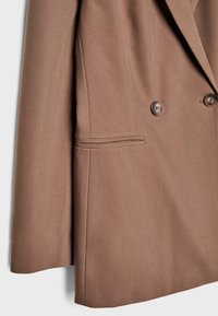 Manche de blazer marron sur mesure et avant inférieur avec deux boutons et poche latérale sur fond blanc.