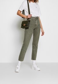 Hoch geschnittene olivgrüne Jeans mit schmalem Schnitt, kombiniert mit einem weißen Rundhalsausschnitt-T-Shirt und weißen Sneakers. Eine kleine schwarze Handtasche setzt einen eleganten Akzent.