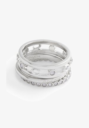 Anillos apilados de plata con varios diseños, que incluyen recortes circulares y piedras preciosas claras, con marca grabada en el interior de las bandas.