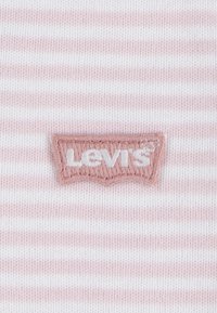 Levi's logotip na pozadini u ružičasto-bijelim prugama, s izvezenim dizajnom i laganom teksturom tkanine.