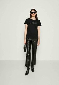 Mulher a usar óculos de sol pretos, t-shirt preta da "GUESS", calças de pele pretas, botas pretas de bico e a segurar uma pequena mala preta.