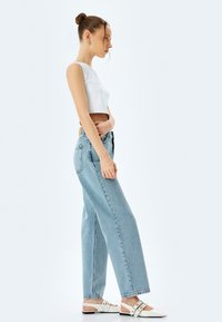 Débardeur blanc raccourci, jeans taille haute bleu clair, et sandales blanches à glisser avec des détails de lanières, vues de côté.
