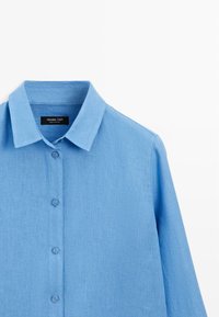Lyseblå langærmet skjorte med knapper og spids krave, lavet af linnedstof, vist på en hvid baggrund.