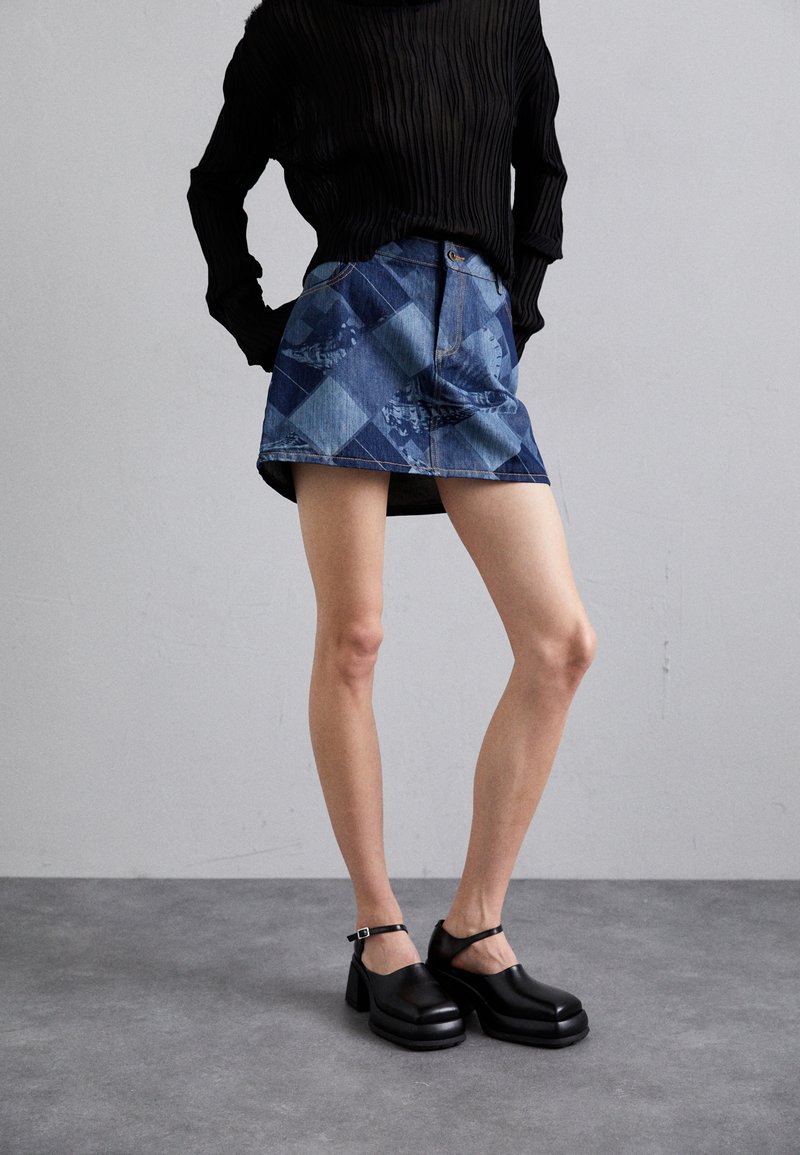 Denim mini krilo s patchwork dizajnom v modrih odtenkih, v kombinaciji s črnimi platformnimi čevlji na peto z gležnjenjem in gladko teksturo.
