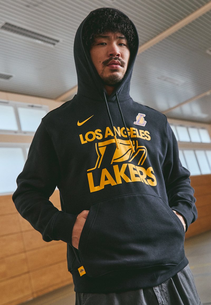 Hoodie preto com "LOS ANGELES LAKERS" em grande fonte amarela e um logótipo dos Lakers. Cordões ajustáveis e bolso canguru frontal.