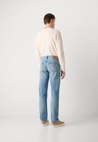 Jeans de denim azul claro con un ajuste relajado, que presentan un parche de logo marrón en la cintura trasera y en los bolsillos traseros. Llevados con un suéter color crema.