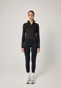 Czarna dopasowana kurtka z wysokim kołnierzem i zamkiem błyskawicznym, zestawiona z czarnymi legginsami i białymi skarpetkami. Modelka nosi wielokolorowe sneakersy.