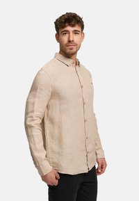 Young man with curly hair wearing a beige button-up long-sleeve shirt and black pants, standing against a plain white background.