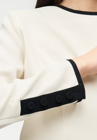 Κρεμ μπλούζα με μαύρες λεπτομέρειες και αντίθετο λαιμόκοψη, με στρογγυλά μαύρα κουμπιά καλυμμένα με ύφασμα στο μανίκι.