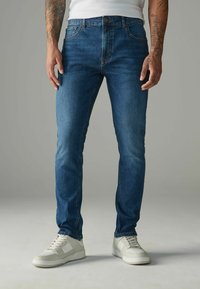Slim-fit džínsy vo farbe tmavomodrej s jemným blednutím, predné vrecká a zapínanie na gombík. V kombinácii s bielymi teniskami.
