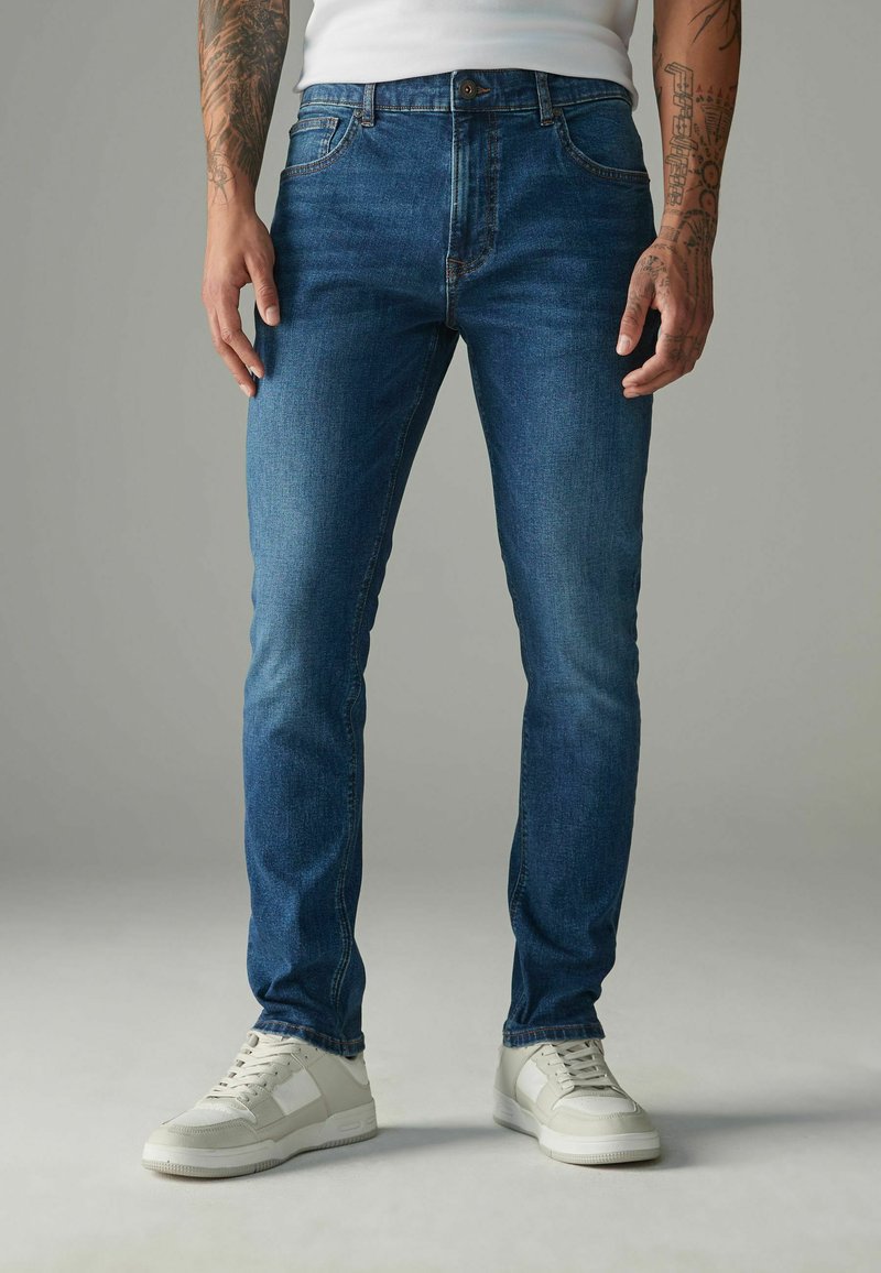 Slim-fit džínsy vo farbe tmavomodrej s jemným blednutím, predné vrecká a zapínanie na gombík. V kombinácii s bielymi teniskami.