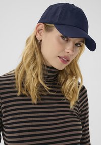 Navy blue cap made of fabric, featuring a curved brim and six-panel design. Worn with a black and brown striped turtleneck top.