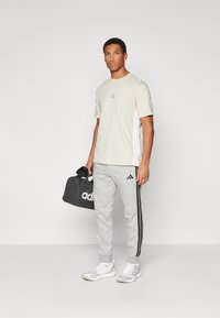 Helles beiges T-Shirt mit kurzen Ärmeln und kleinem Logo, graue Jogginghose mit schwarzen Seitenstreifen und schwarze Sporttasche mit weißer Markenkennzeichnung.