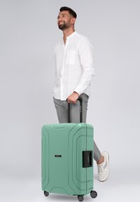 Uomo in camicia bianca e pantaloni grigi in piedi con una valigia rigida verde menta con manico estensibile e ruote, sorridente e che guarda in alto.